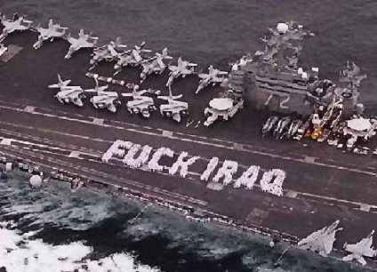 Photo of the deck of an aircraft carrier, taken from one or two hundred feet above as it plows through the ocean water. Many white aircraft with folded wings line the deck. A large group of sailors and/or pilots wearing white uniforms are standing on the angled runway of the deck, having formed themselves into two large white words which read: FUCK IRAQ.